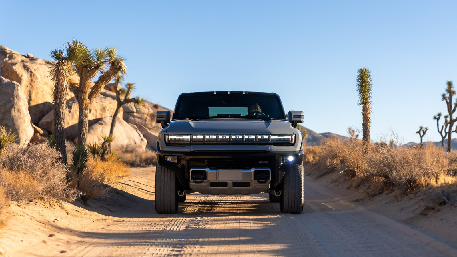 A large black SUV drives on a desert dirt road, surrounded by rocks and sparse vegetation under a clear blue sky.