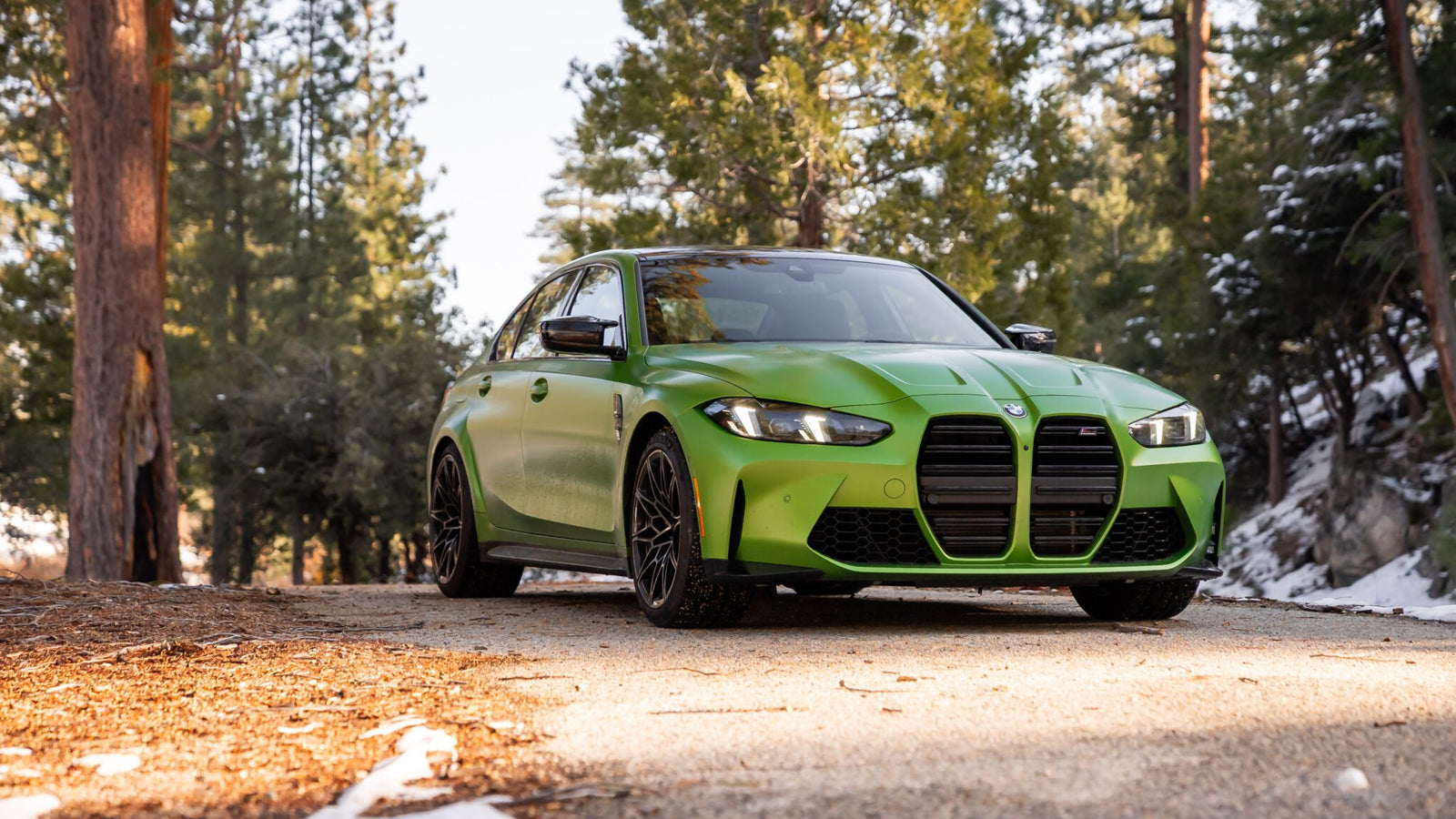 A green BMW sedan is parked on a forested road with trees and patches of snow in the background.