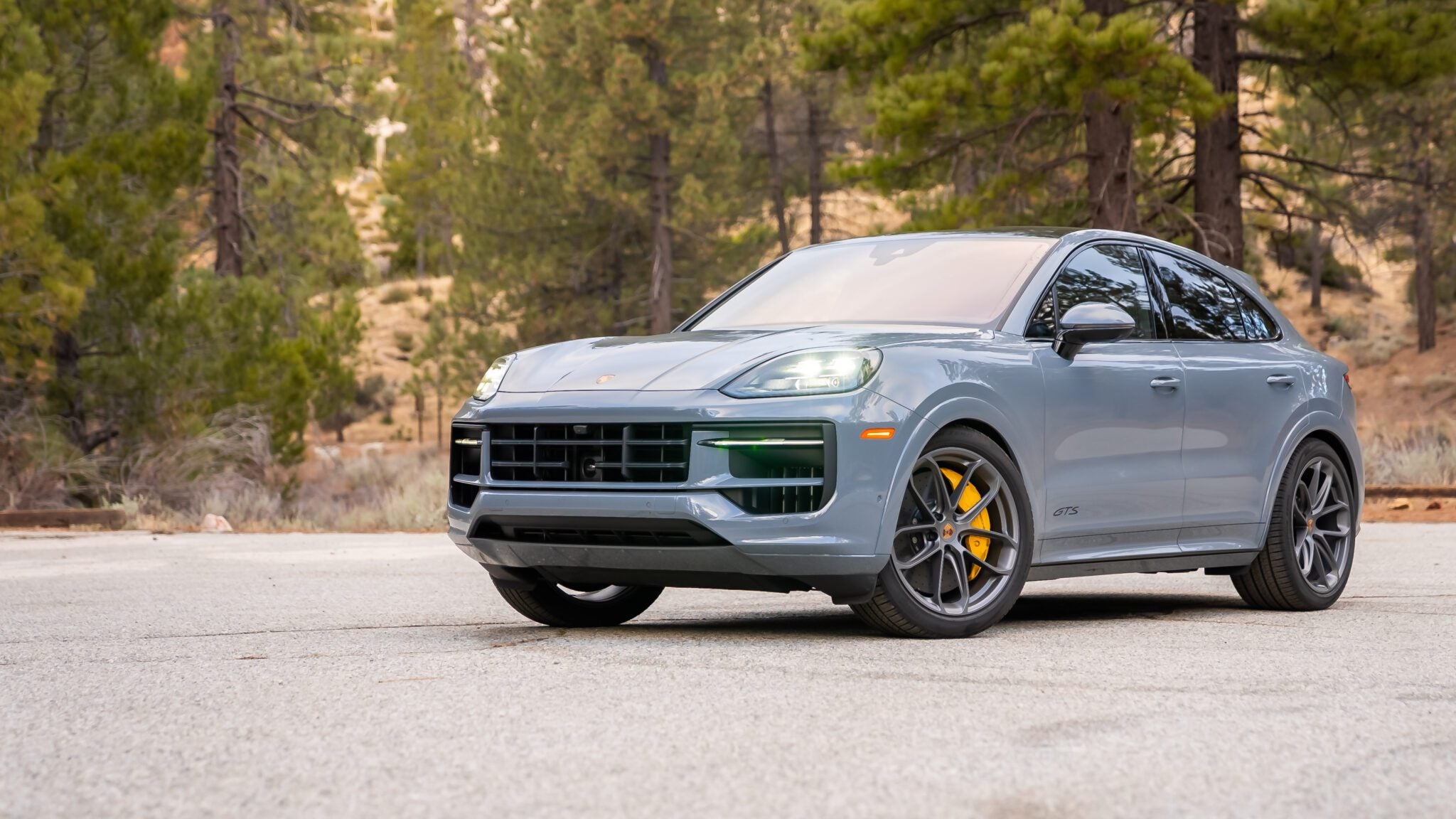 A gray Porsche SUV with yellow brake calipers is parked on a paved surface, surrounded by trees and a forested background.