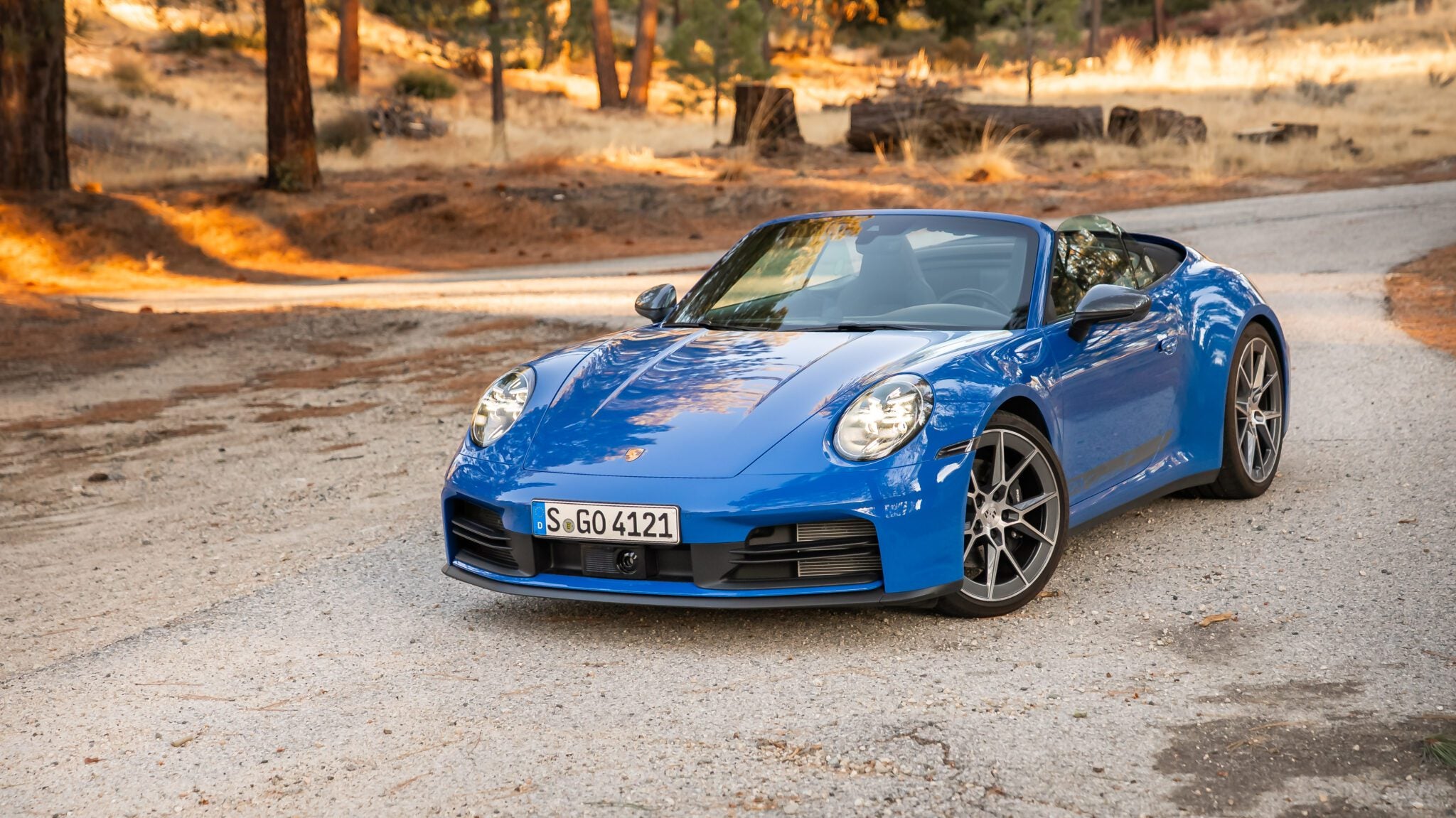 A blue convertible sports car is parked on a gravel road in a wooded, dry landscape with scattered trees and sunlight.