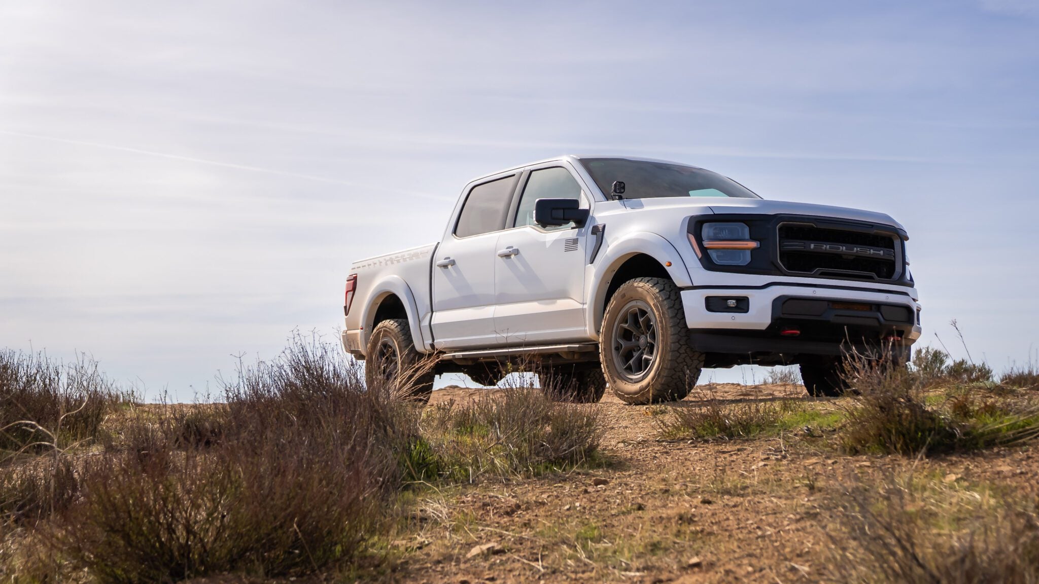 A white pickup truck parked on a dirt road, surrounded by dry grass and bushes under a clear sky.