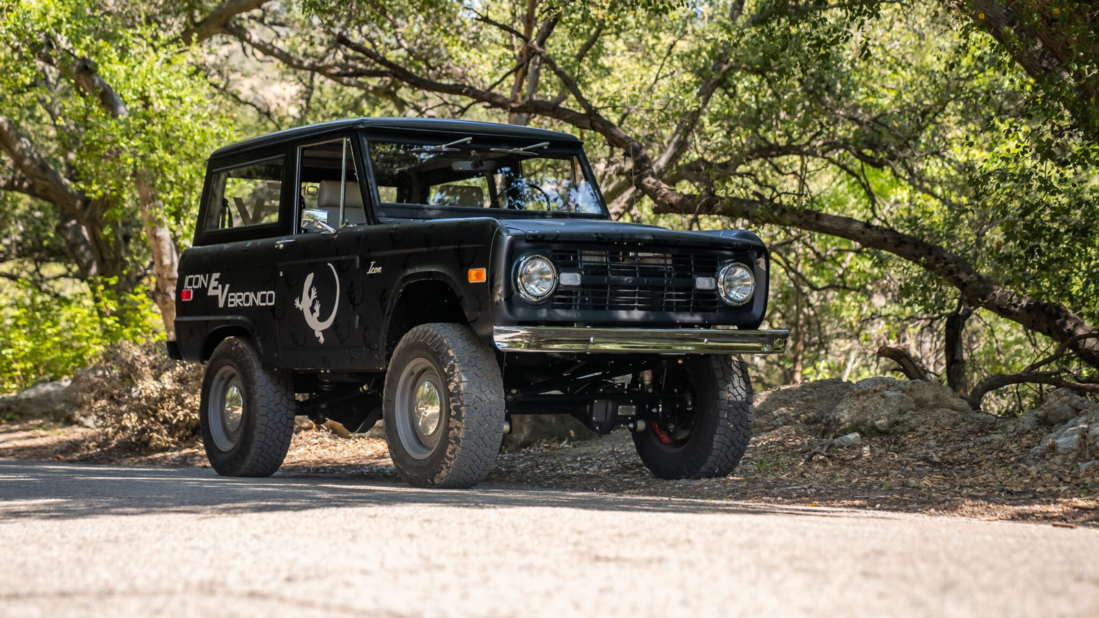 A vintage black off-road vehicle is parked on a forested road, surrounded by trees and dappled sunlight.