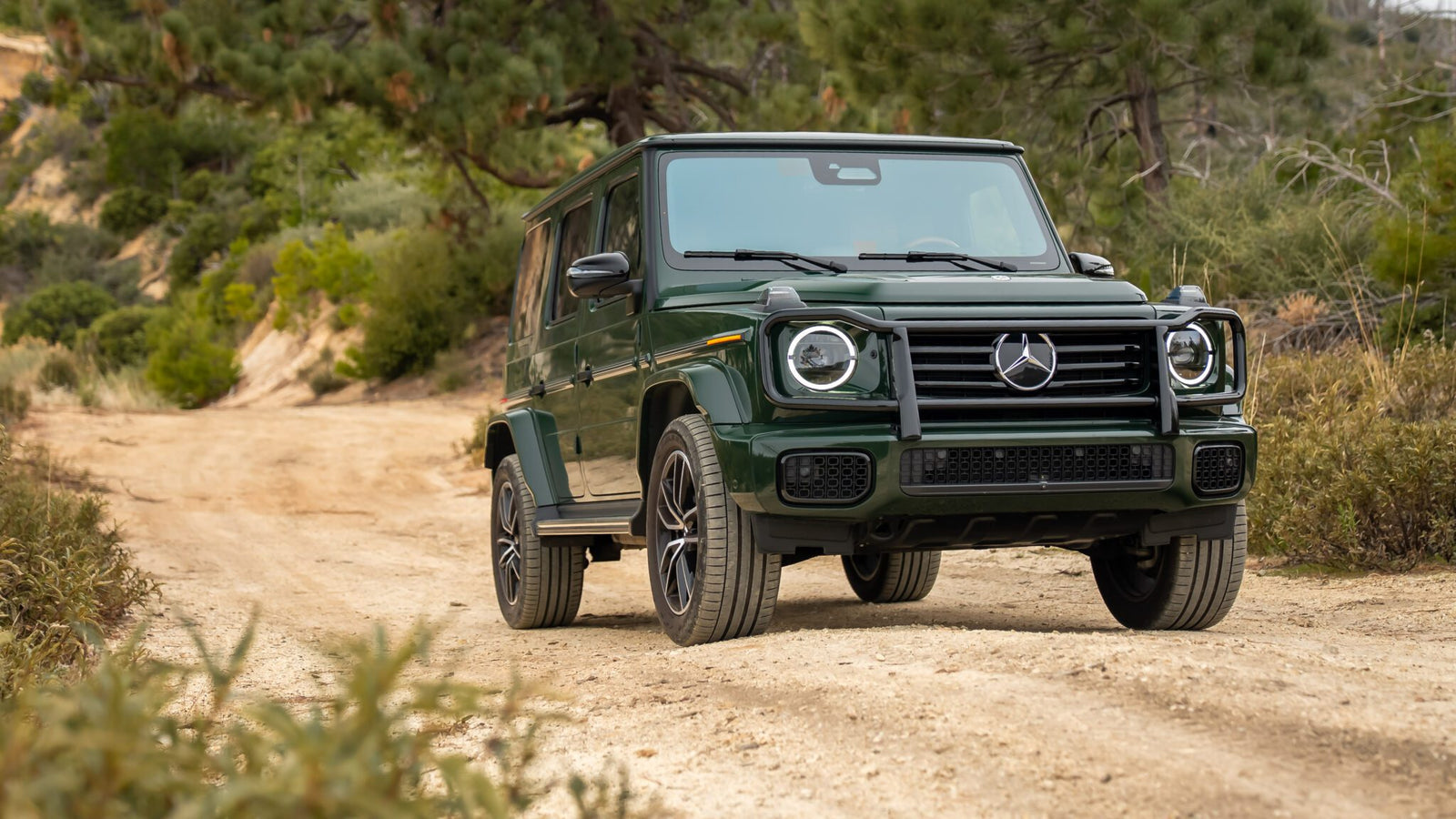 A green Mercedes-Benz G-Class SUV is parked on a dirt trail surrounded by trees and shrubs in a natural outdoor setting.