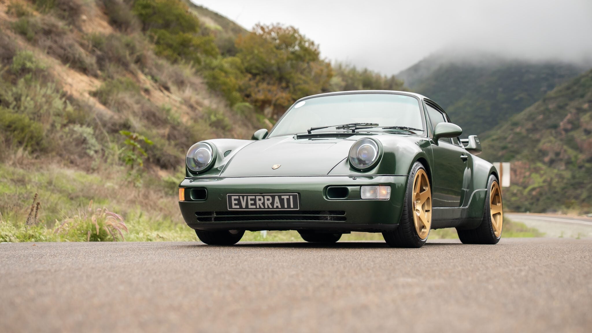 Green sports car with a front license plate reading "EVERCAT" parked on a scenic, winding road surrounded by hills and cloudy sky.