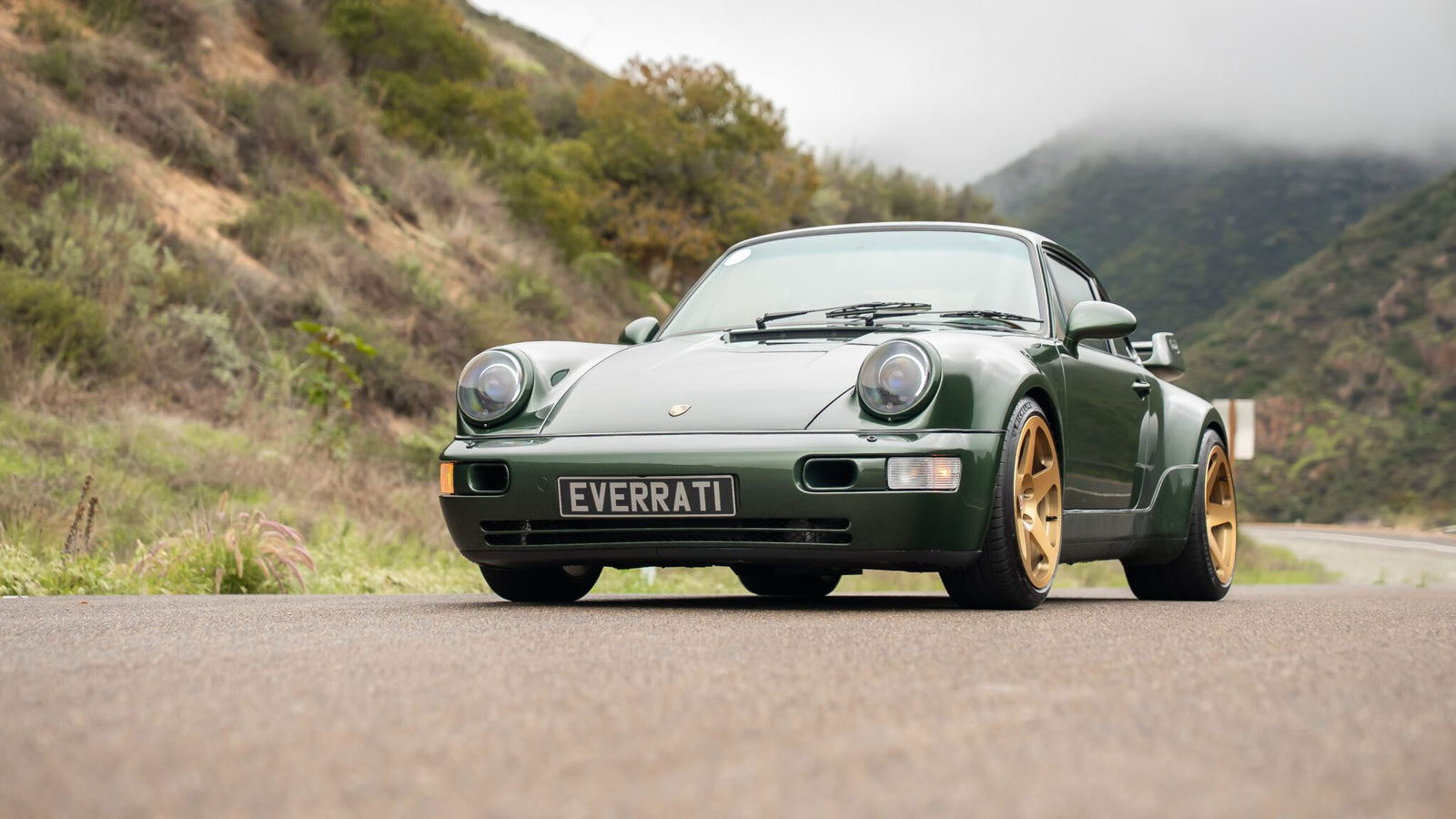 Green sports car with a front license plate reading "EVERCAT" parked on a scenic, winding road surrounded by hills and cloudy sky.