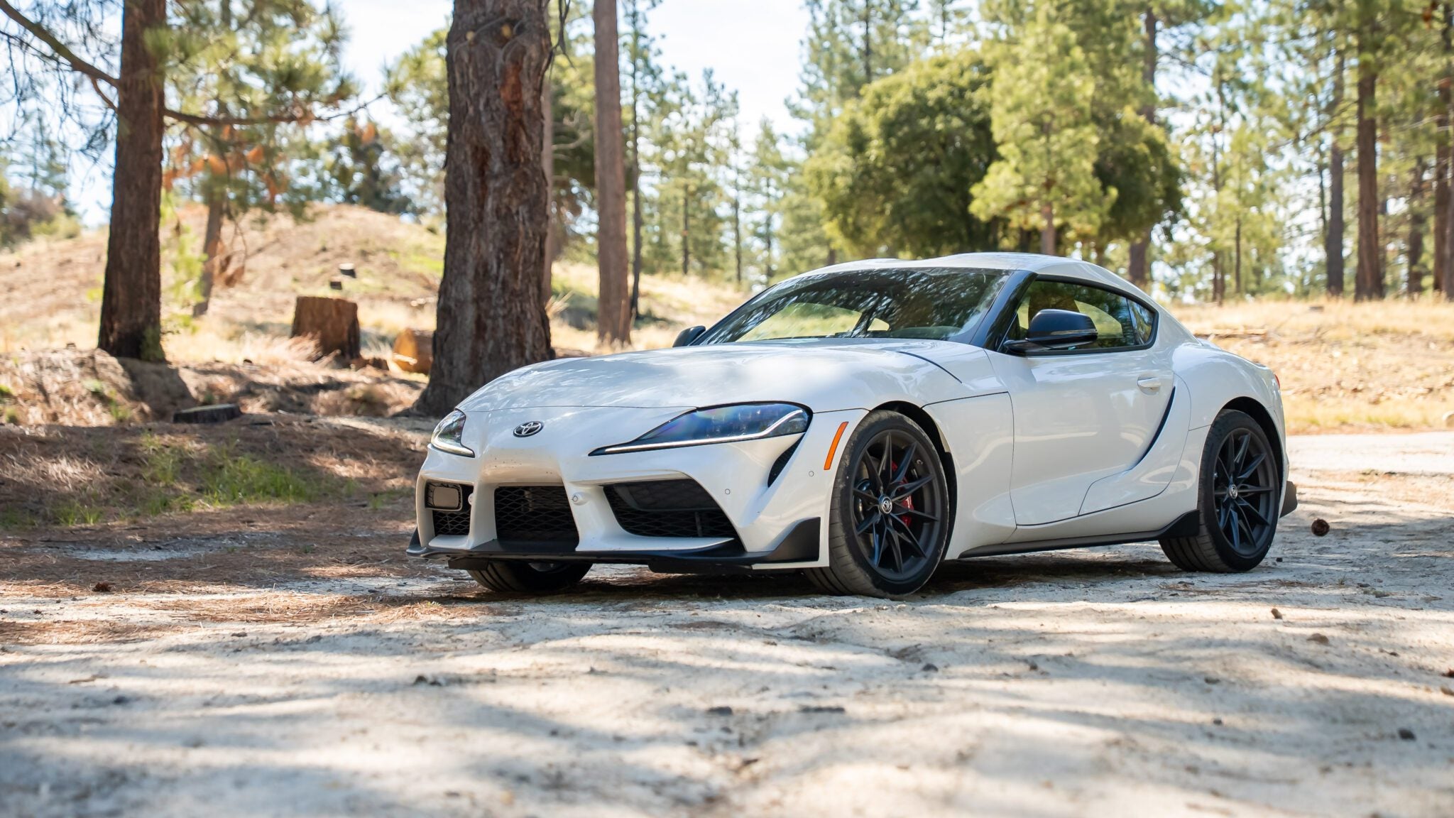 A white Toyota Supra sports car is parked on a dirt road surrounded by trees and natural landscape.