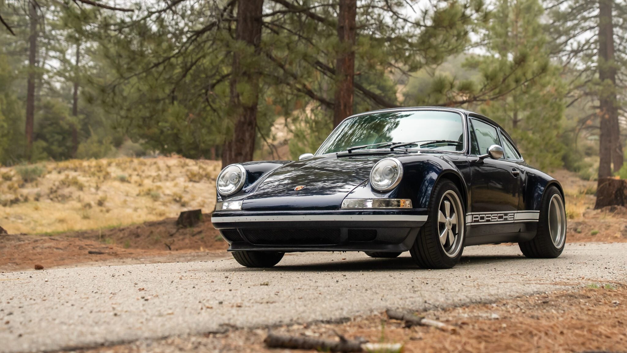 A classic dark blue Porsche 911 is parked on a forest road, surrounded by trees and natural scenery.