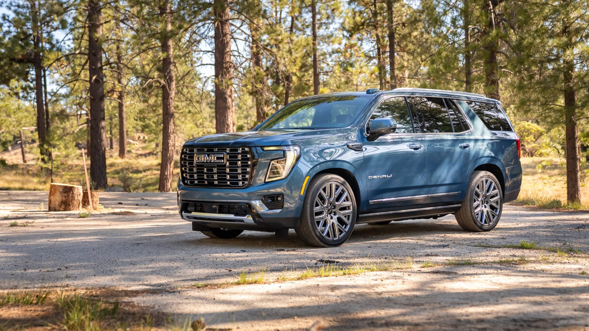 A blue GMC Yukon Denali SUV is parked on a dirt road in a wooded area with tall pine trees in the background.