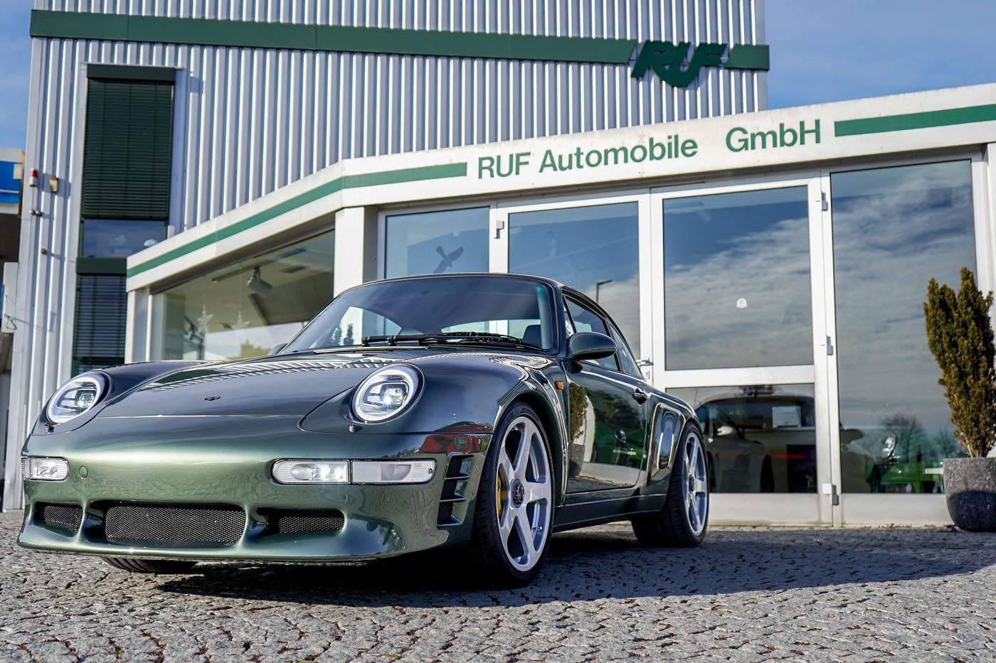 A sleek green 993 Porsche 911 is parked in front of a building with RUF Automobile GmbH written on it, its presence enhanced by modern LED lighting.