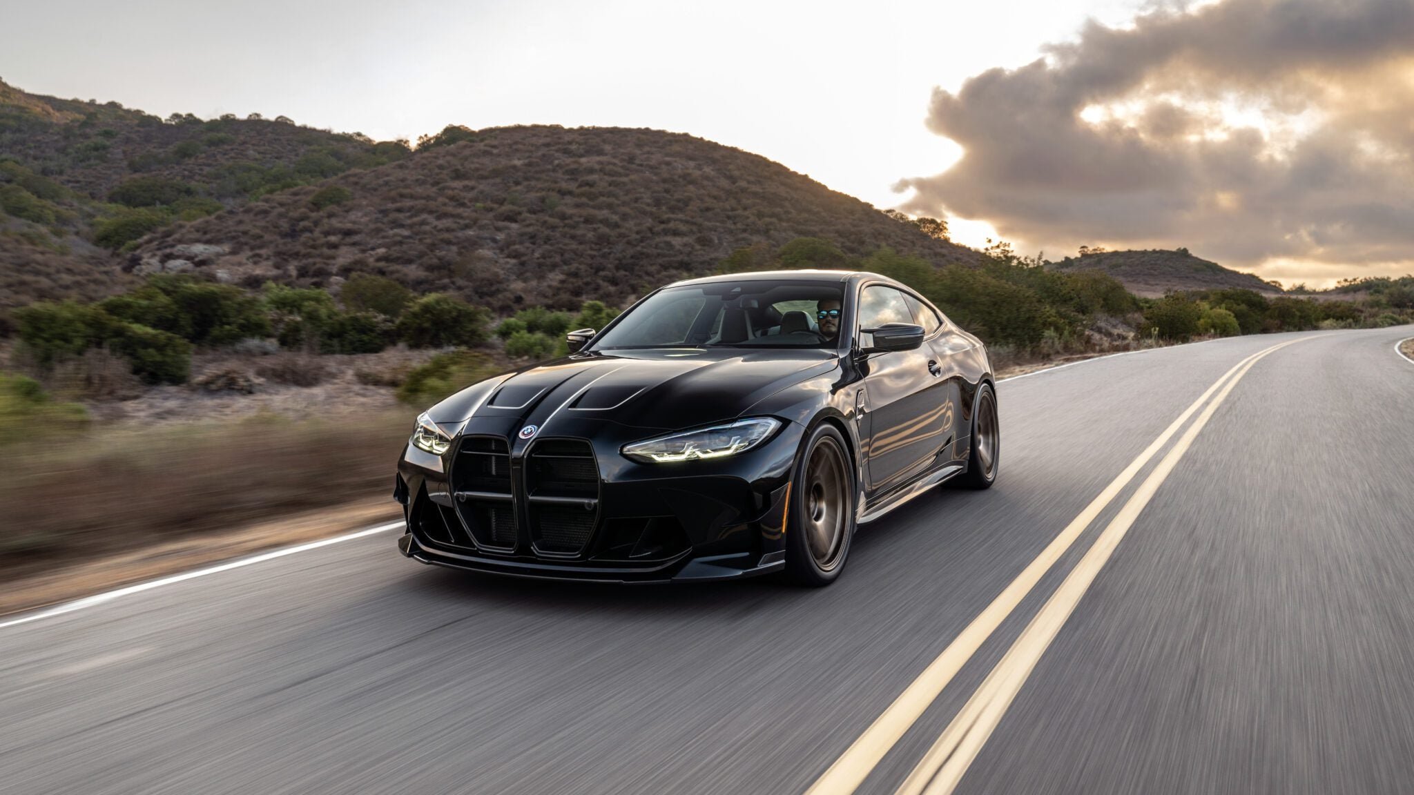 A black sports car drives on a scenic, empty road surrounded by hills and a cloudy sky.