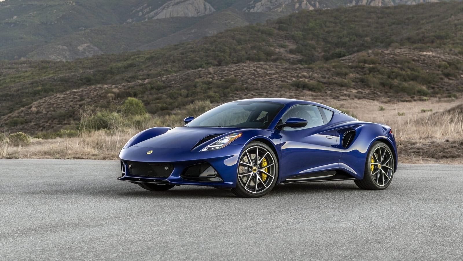 A blue sports car is parked on a road with mountains in the background.