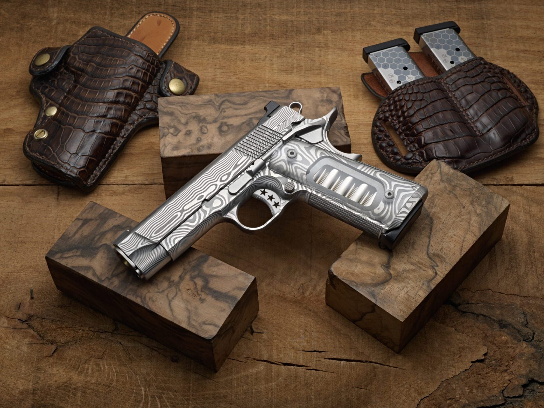 The Ultimate Carry 1911 by Cabot Guns is a masterpiece of American craftsmanship, featuring a silver-patterned pistol with wooden grips elegantly displayed on wooden blocks, surrounded by leather holsters and magazine pouches.