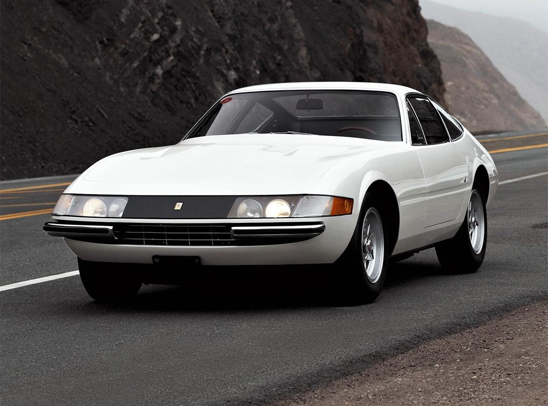 A white 365 GTB/4 Daytona classic sports car is parked on a winding mountain road with rocky cliffs in the background, exuding timeless elegance reminiscent of Ferrari Beverly Hills.