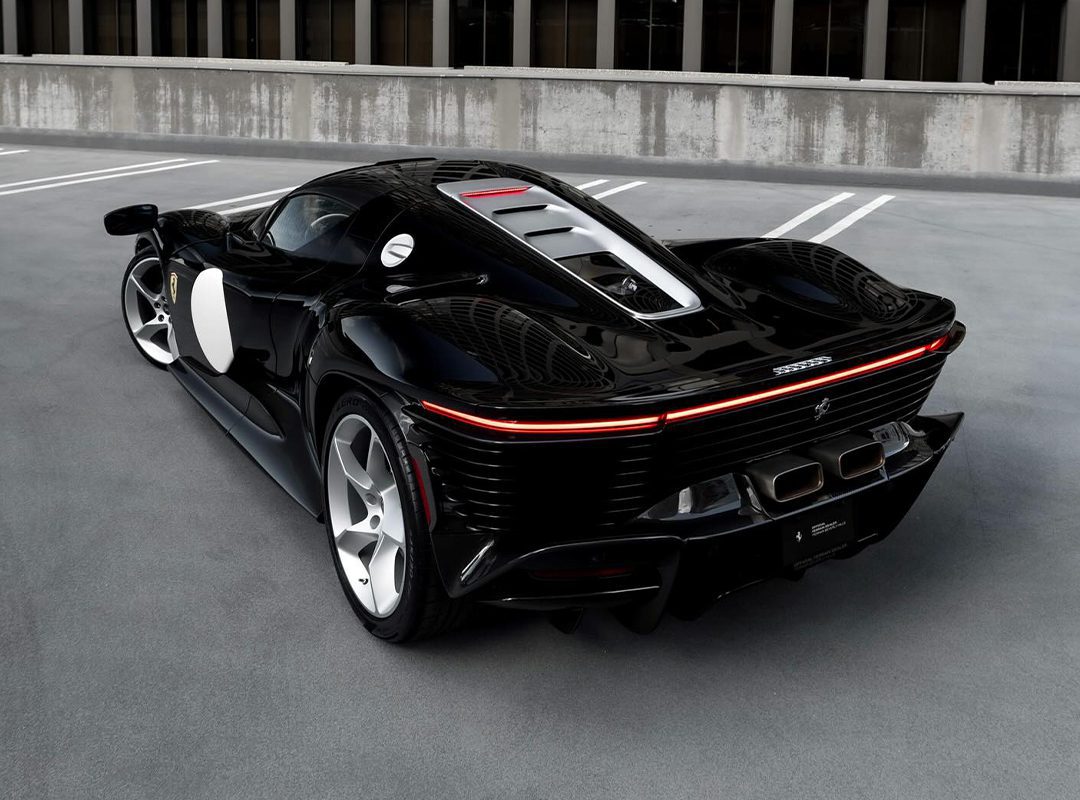 A sleek, black Ferrari Daytona SP3 sports car is parked on a Beverly Hills rooftop with a modern building in the background. The car boasts a low profile, aerodynamic design, and distinctive rear lights.