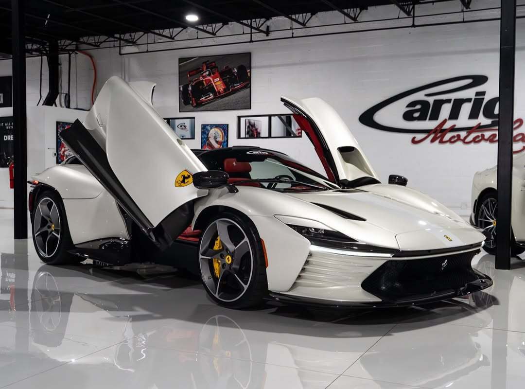 A white Ferrari Daytona SP3 with butterfly doors open is displayed in a showroom with glossy floors and automotive decor—a stunning 2023 Ferrari for sale at Carrior Motor Cars.