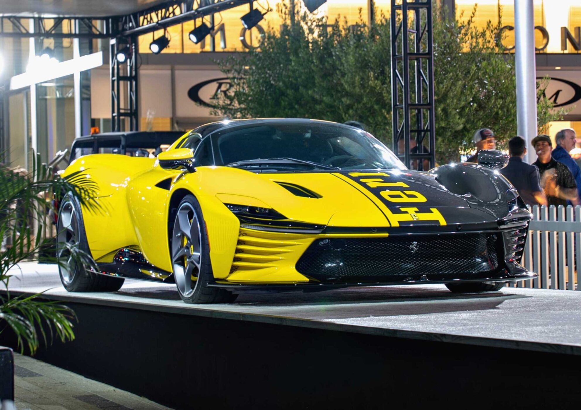 A yellow and black Ferrari Daytona SP3, worth $26 million, is displayed on an outdoor platform at a car show, drawing the attention of billionaire car enthusiasts amid people and lights in the background.