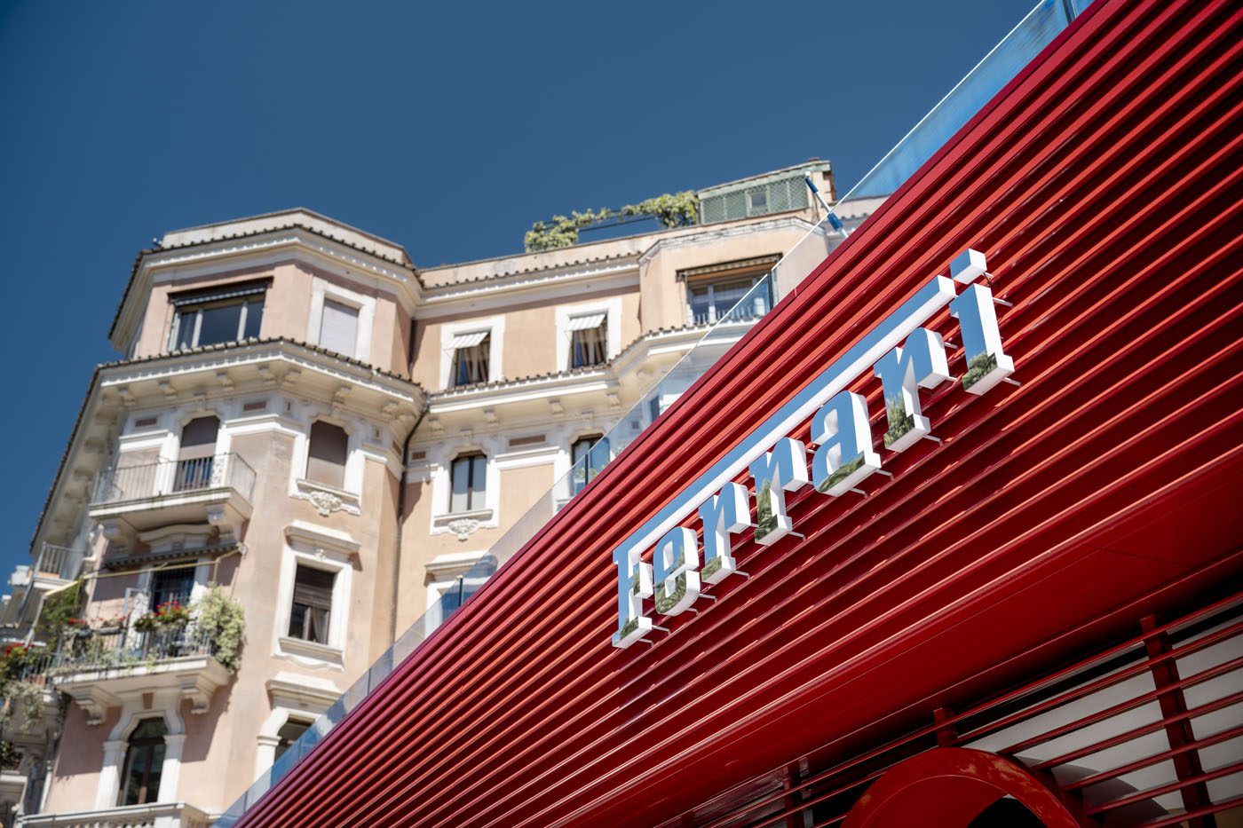 A red Ferrari storefront with sleek dealer corporate identity features a modern design in front of a beige, multi-story historic building under a clear blue sky.