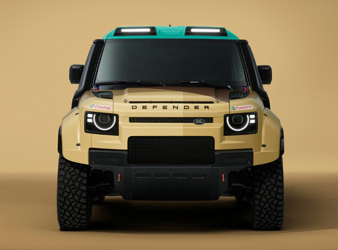 Front view of a tan Land Rover Defender with rugged tires, black accents, a green roof, and Castrol sponsor stickers, set against a beige background.