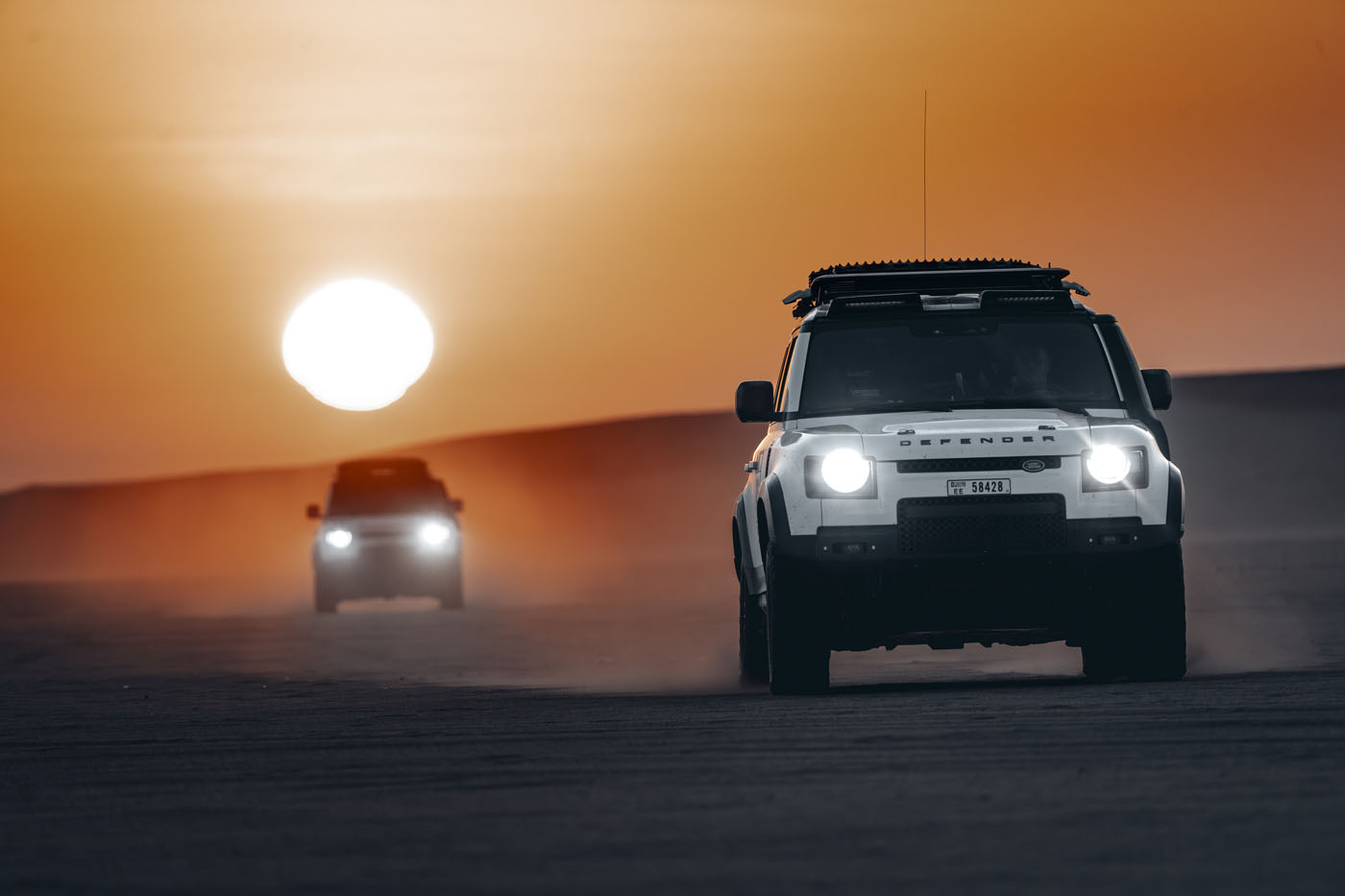 Two Defender vehicles race across sandy terrain at sunset, dust trailing behind as they follow their route planning, evoking the thrill of the Dakar Rally.