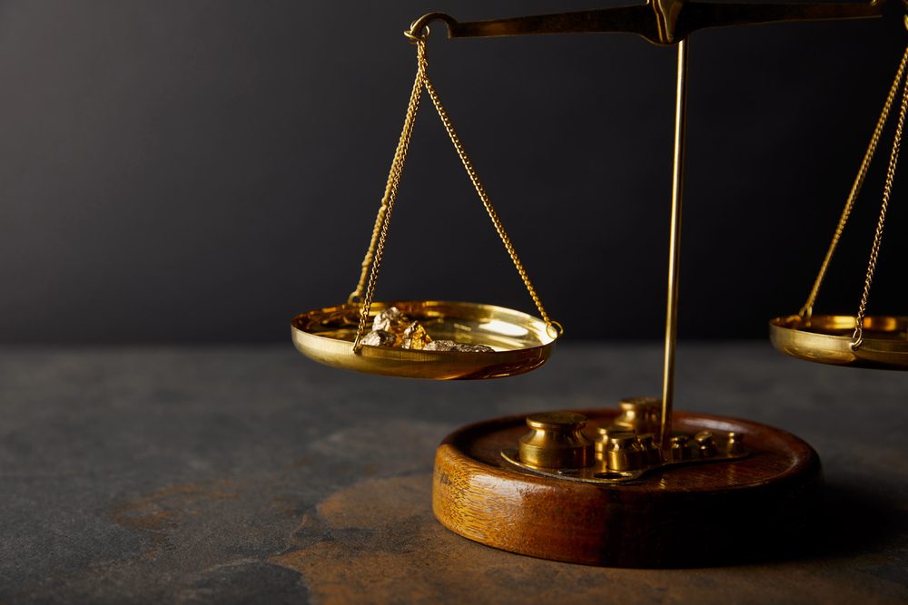 A gold balance scale holds gold nuggets on one side and weights on the other, symbolizing asset valuation, placed on a dark stone surface with a black background.