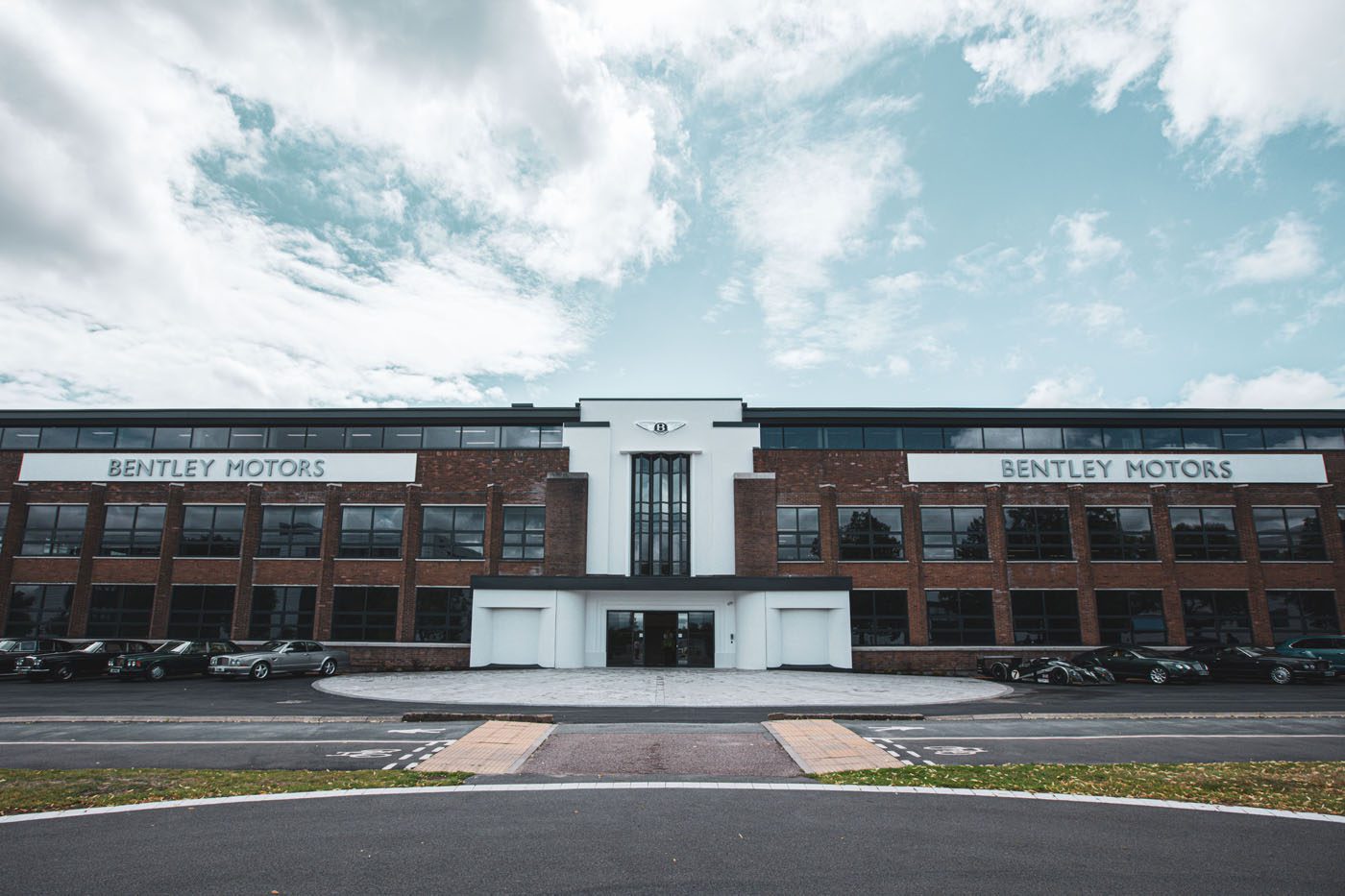 A large brick and glass building with "Bentley Motors" signs on both sides, several cars parked in front, and a partly cloudy sky above.