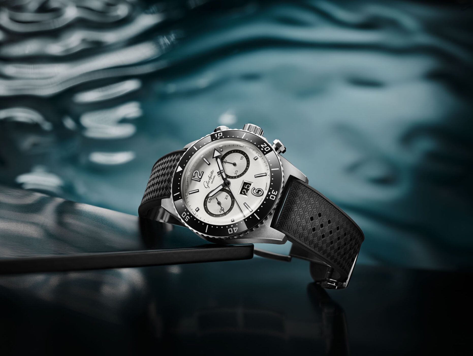 A new Chronograph wristwatch with a silver face and black strap rests elegantly on a reflective dark surface, complemented by its watery background.