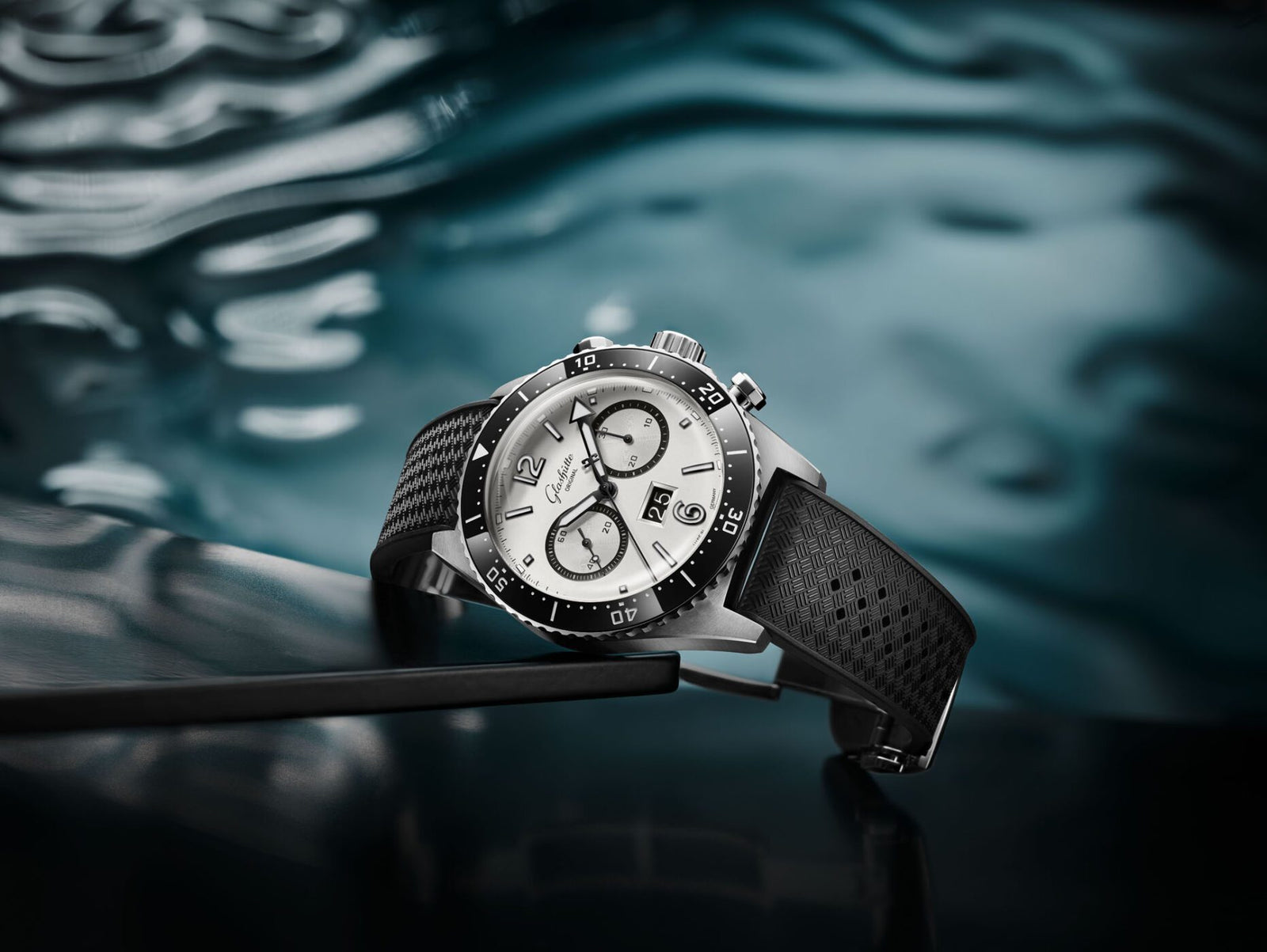 A new Chronograph wristwatch with a silver face and black strap rests elegantly on a reflective dark surface, complemented by its watery background.