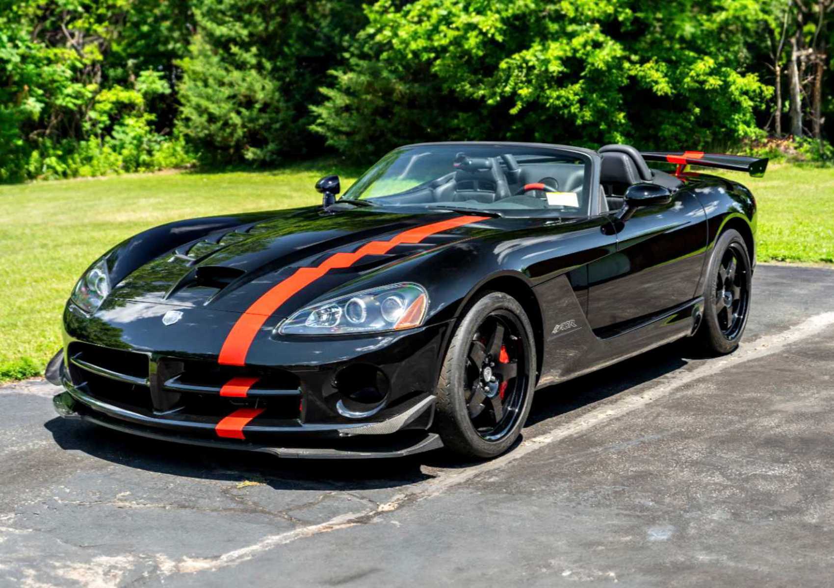 A black convertible sports car with red racing stripes and a rear spoiler is parked on a paved driveway with greenery in the background. Bid On This 2010 Dodge Viper SRT-10 Roadster ACR With Just 20 Miles today!.