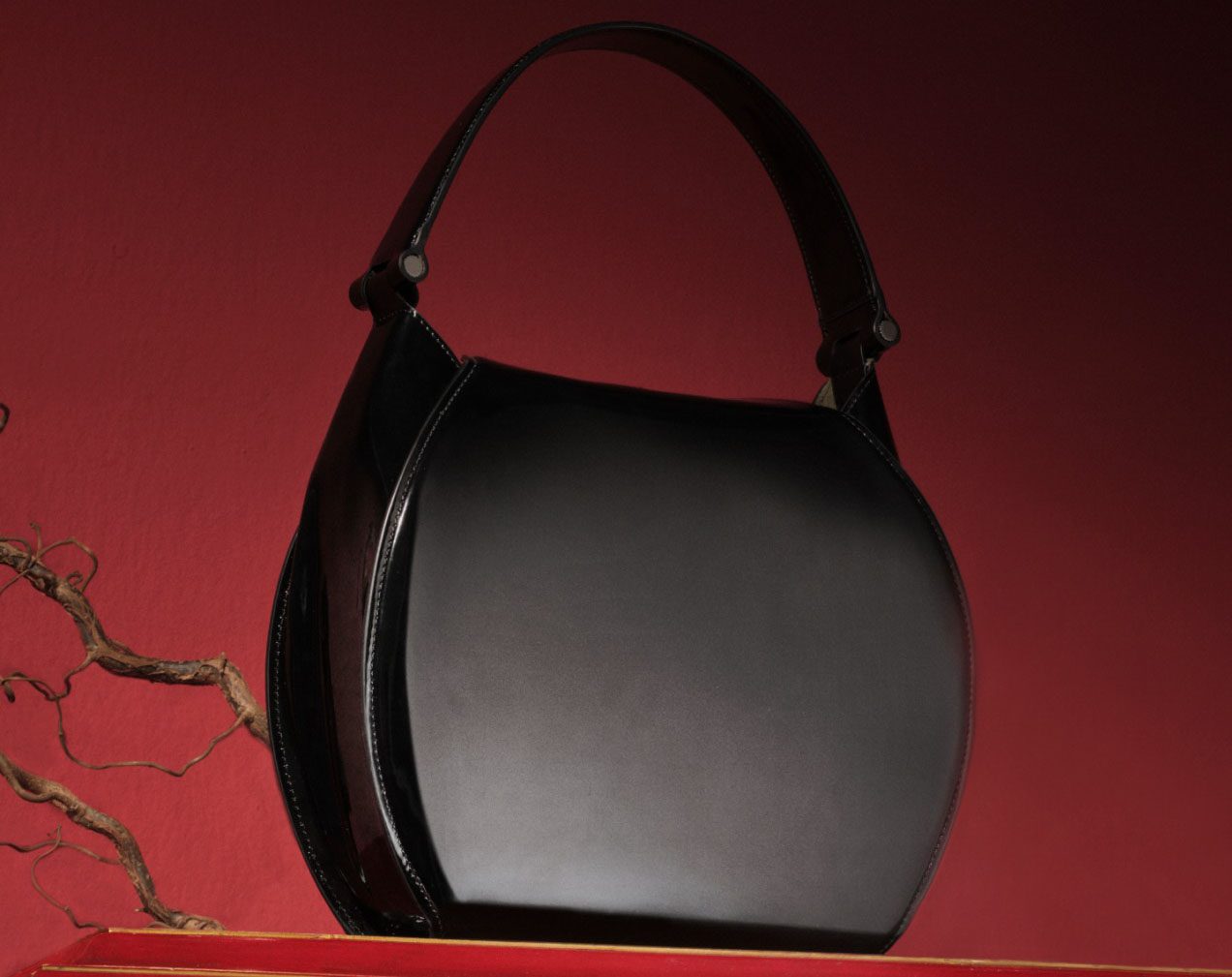 A black leather Ferrari handbag with a curved design sits strikingly against a red background, reminiscent of the elegant curves of the Dino 246, with a dry branch positioned on the left side.