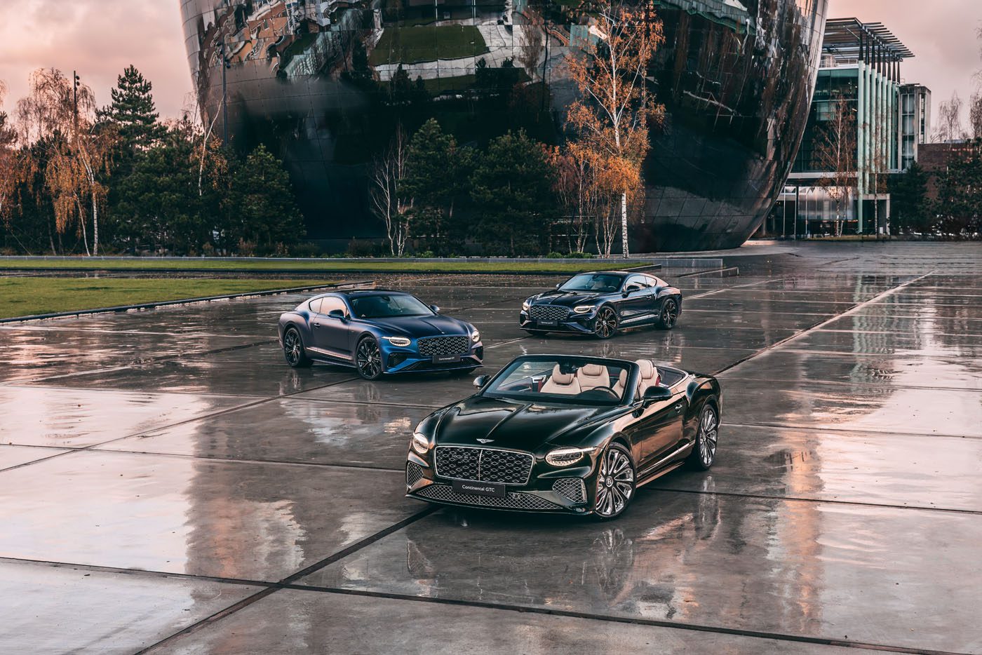 Three luxury Bentley cars—one black convertible in the front and two black coupes behind, part of the exclusive Mulliner Collection—are parked on a wet, reflective plaza with modern buildings and trees in the background.