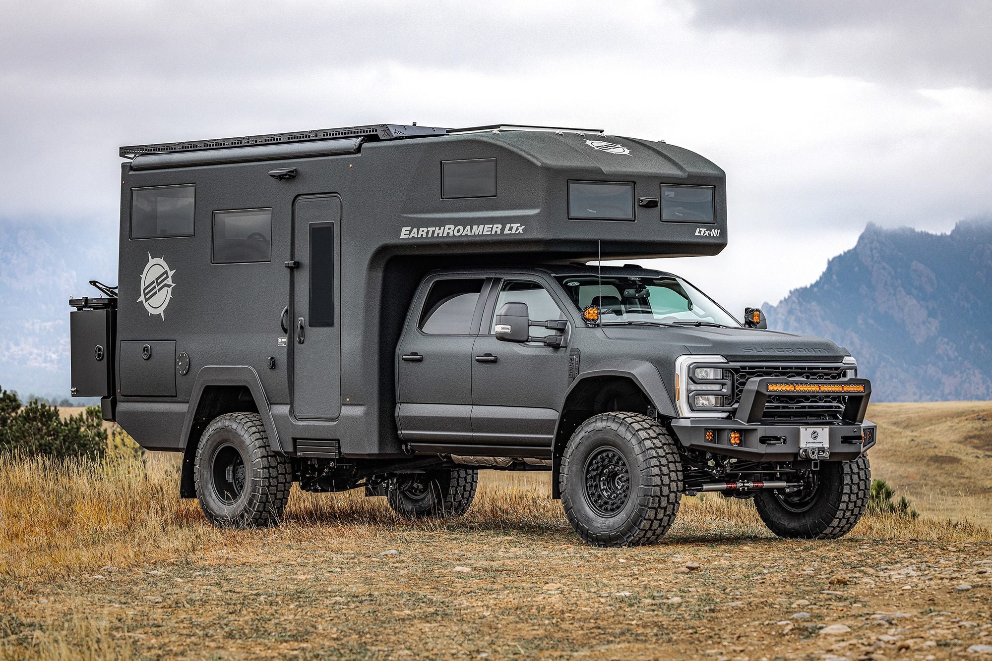 A rugged gray camper truck with off-road tires, the EarthRoamer LTi, is parked in a grassy, mountainous area. This adventure-ready vehicle might just be a highlight when it rolls into the Barrett-Jackson event in Scottsdale 2025.