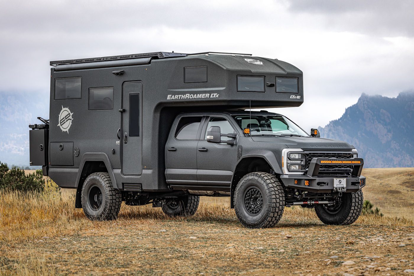 The ultimate expedition vehicle, an EarthRoamer with large tires and a rugged exterior, is parked on grassy terrain with majestic mountains in the background.