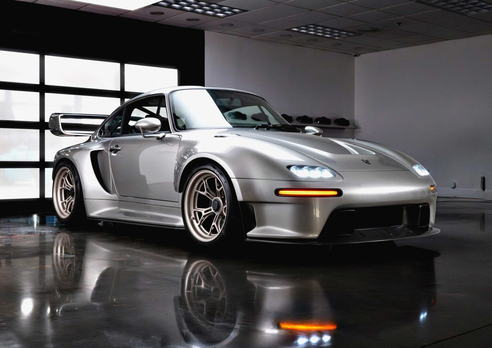 A silver sports car with a wide body kit and rear wing, inspired by the Gunther Werks Unveils Limited Edition 1000-HP Slantnose-Inspired F-26 at The Quail, sits indoors on a reflective floor under ceiling lights, headlights aglow.