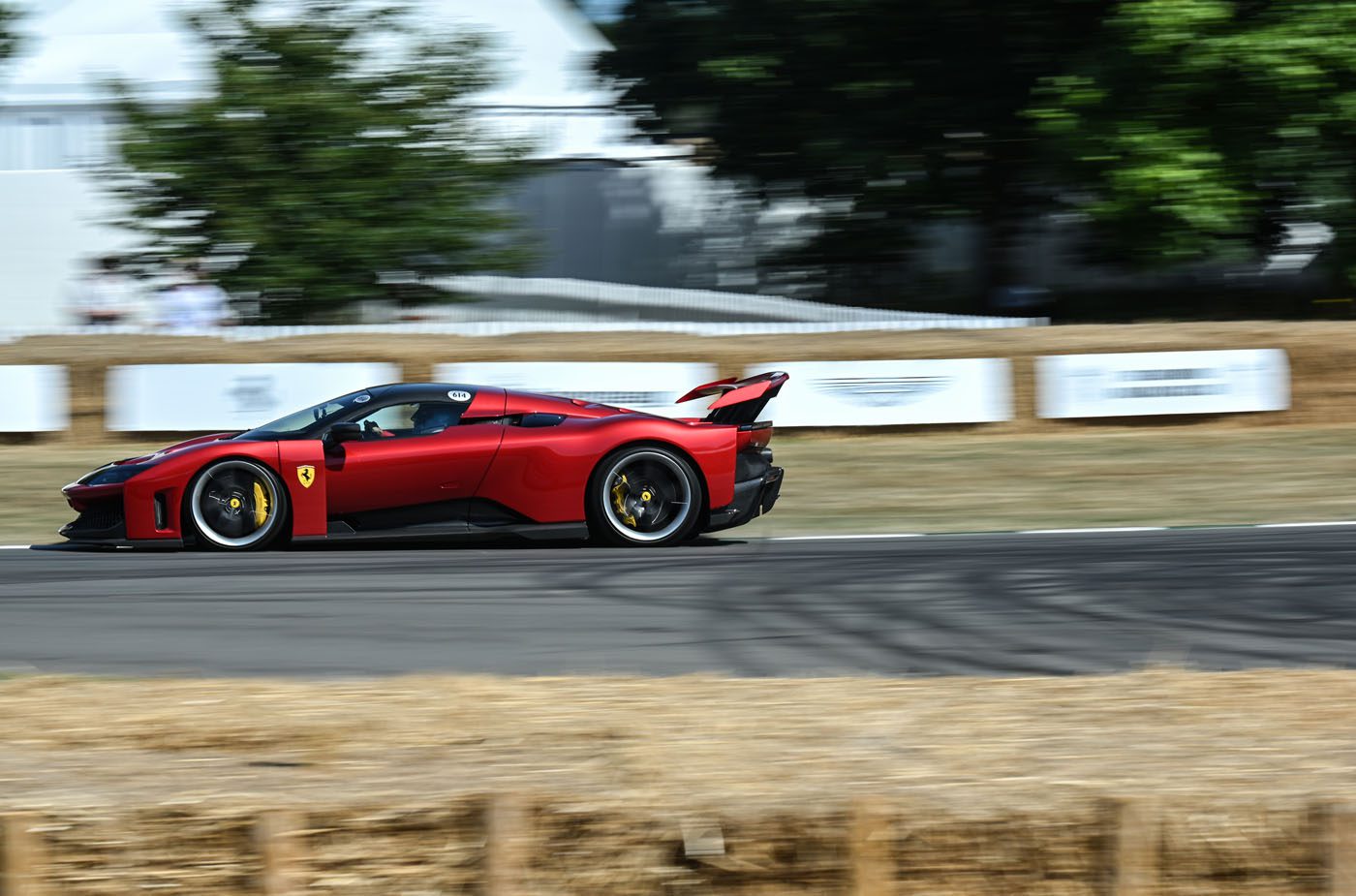 A red Ferrari speeds along the Goodwood racetrack, bordered by hay bales, with trees and white fencing in the background.
