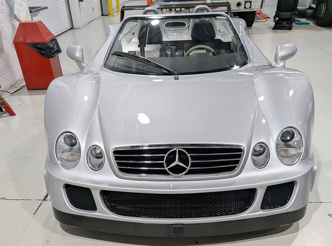 A silver Mercedes-Benz CLK-GTR Roadster convertible sports car, valued as a $13M car, is parked indoors on a clean, white floor with its top down and front view visible.