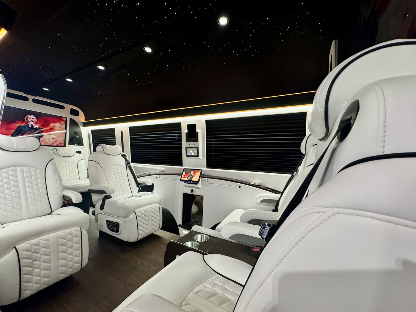 The 2024 Jet Sprinter by First Class Customs features a lavish interior with white leather seats, wood flooring, ambient lighting, and a ceiling that mimics a starry night. A sleek TV screen graces the background, creating an atmosphere of pure luxury.