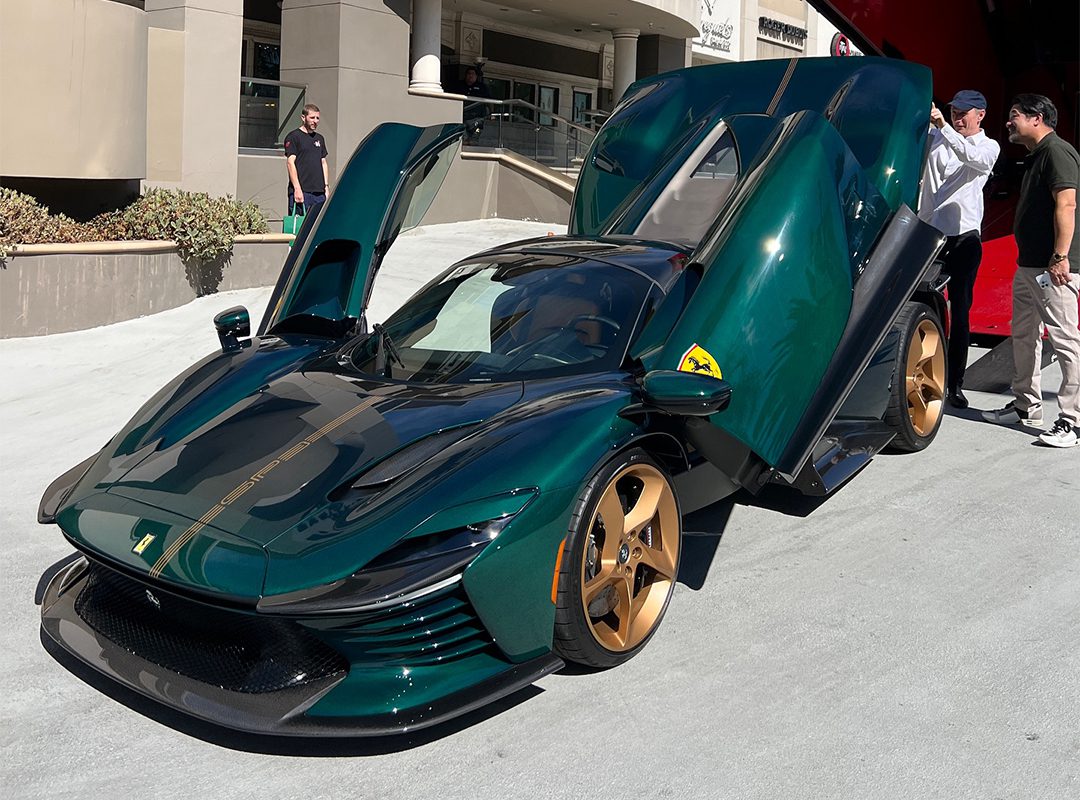 A dark green Daytona SP3 sports car with gold wheels and butterfly doors open, parked on a street with two people nearby, captivates the eye of a true Ferrari collector.