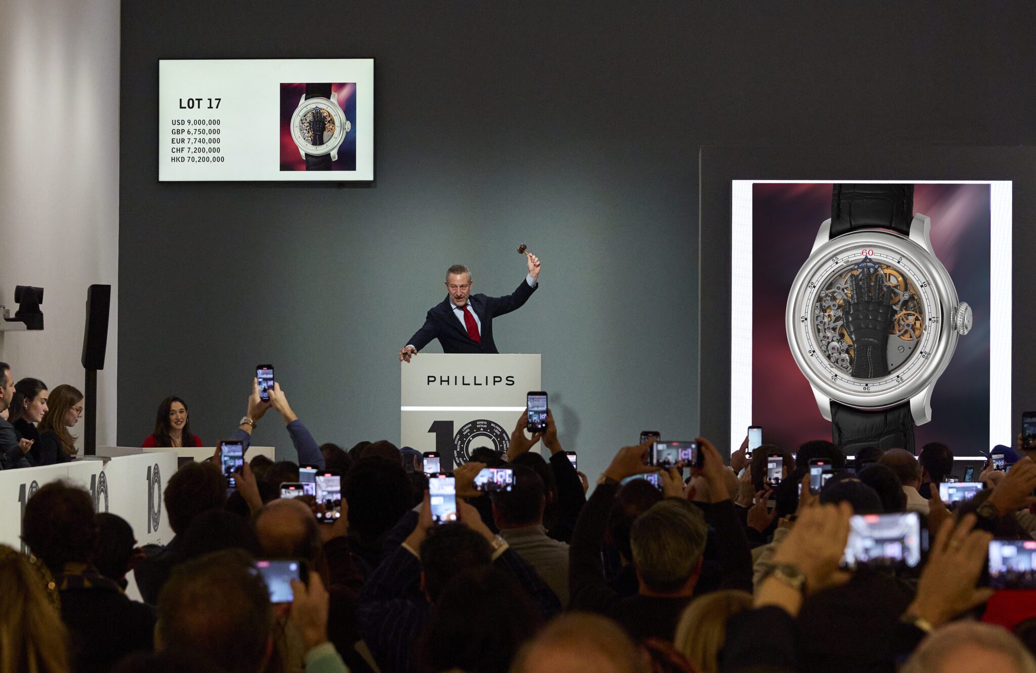 An auctioneer holds a gavel at a Phillips auction, with images and details of a wristwatch displayed on screens and attendees taking photos with phones.