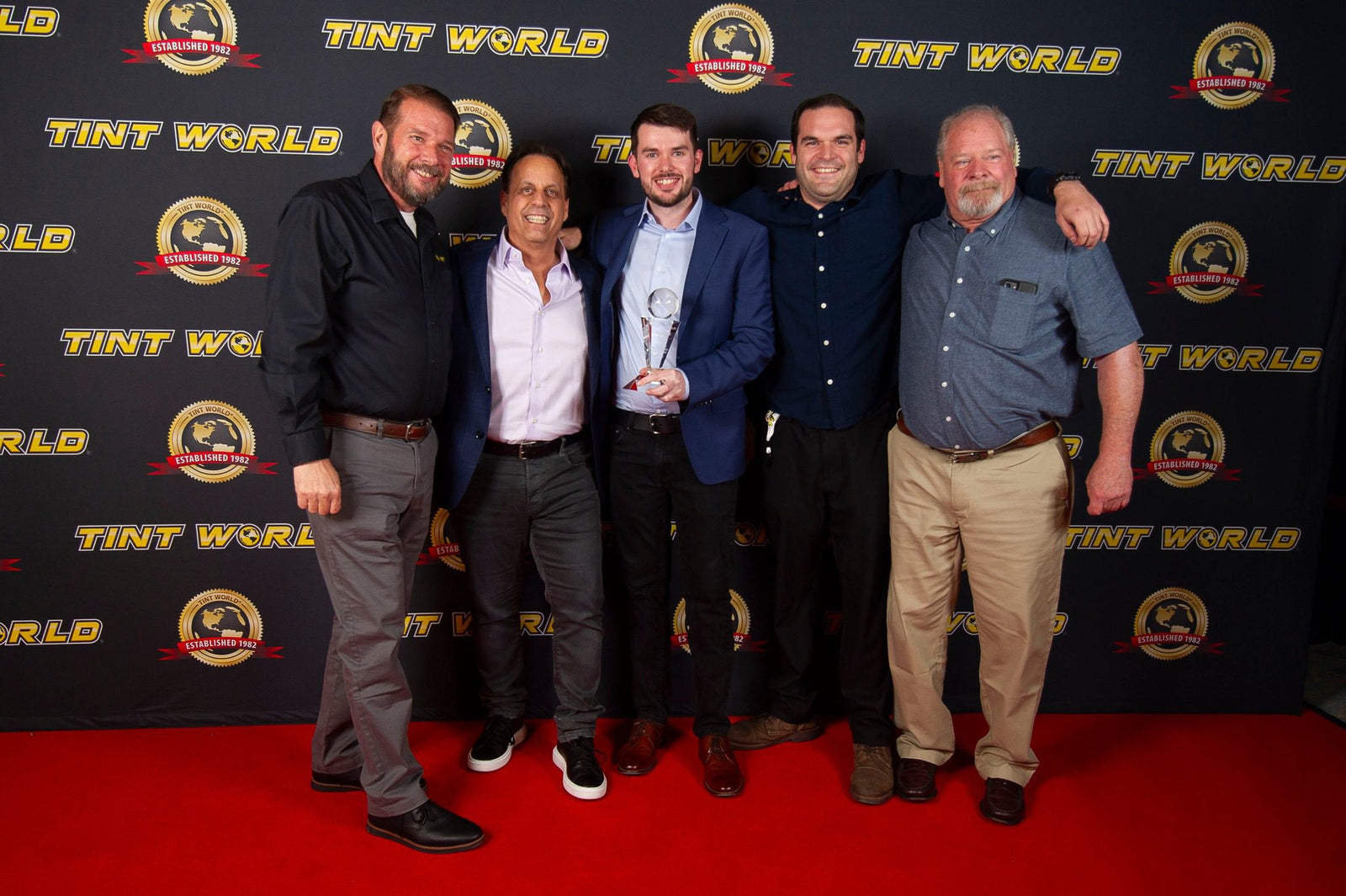 Five men stand proudly on the red carpet, posing for a group photo before a "TINT WORLD" backdrop. One man holds an award, celebrating their status as top-performing franchisees in Tint World®, highlighting their million-dollar achievements with pride.
