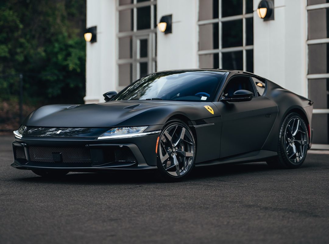 A matte black Ferrari 12Cilindri, boasting 819HP, is parked in front of a white building with large windows and exterior lights—a stunning showcase from Miller Motorcars.