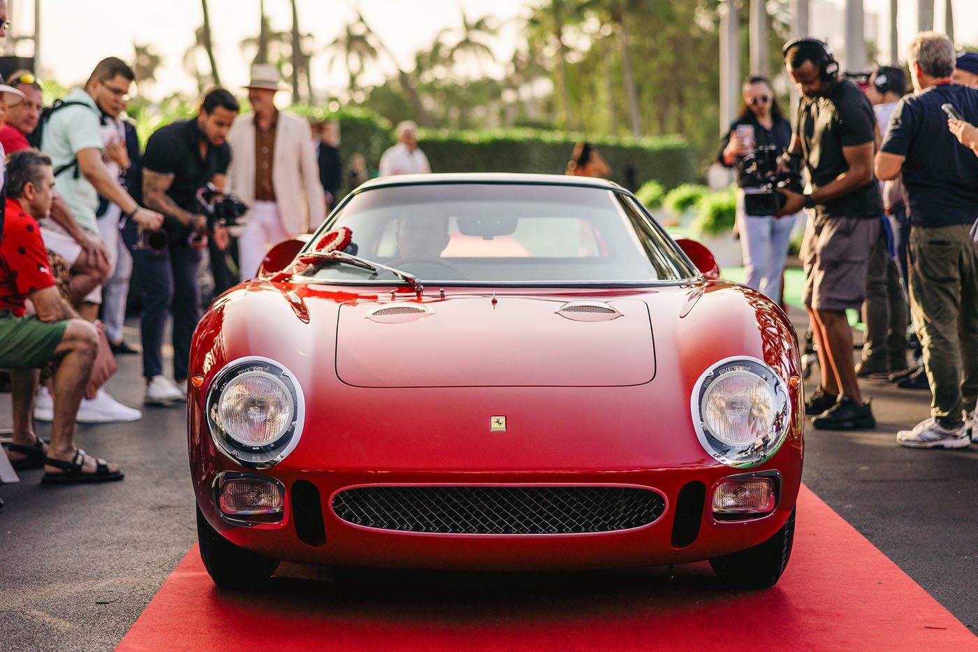 A red vintage Ferrari 250 LM sports car, often hailed as the Best of the Best, is parked on a red carpet, surrounded by a crowd of people snapping photos at the prestigious Cavallino Classic event.