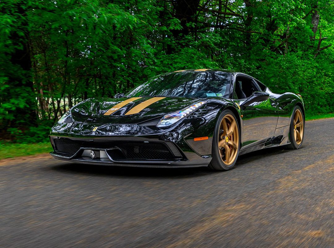 A black Ferrari 458 Speciale with gold wheels and racing stripes drives on a paved road surrounded by green trees, reminiscent of the sleek models seen at the Saratoga Motorcar Auction.