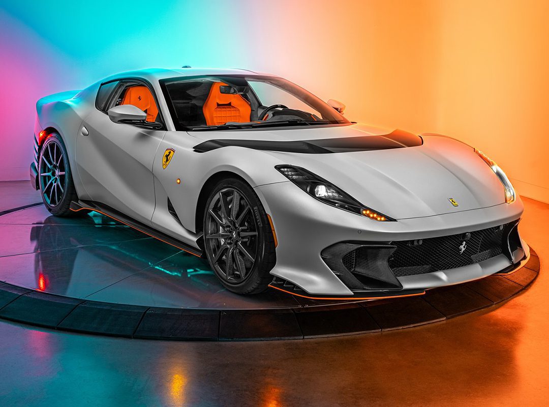 A silver Ferrari sports car with orange interior, part of our certified pre-owned Ferraris collection, is displayed on a rotating platform against a colorful orange and blue gradient background.