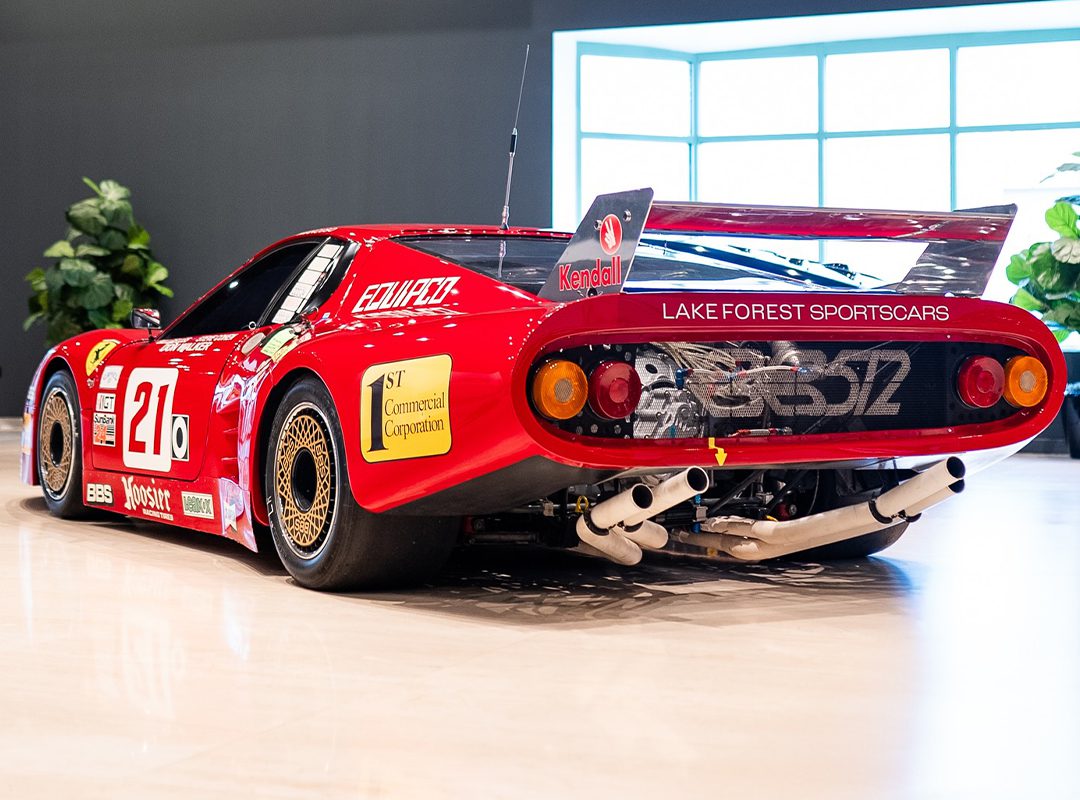 A red vintage Ferrari race car, proudly displaying number 21, with sponsor decals and a large rear spoiler. This 512 BB LM is parked indoors on a light floor, framed by plants and an inviting window backdrop – truly race-ready elegance.