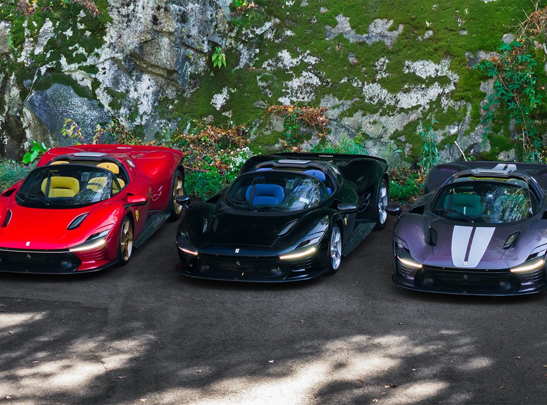 Three sports cars—one red, one black, and an ultra-rare Ferrari Daytona SP3 with white stripes—are parked side by side on a paved area next to a mossy rock wall, possibly showcased by Miller Motorcars.