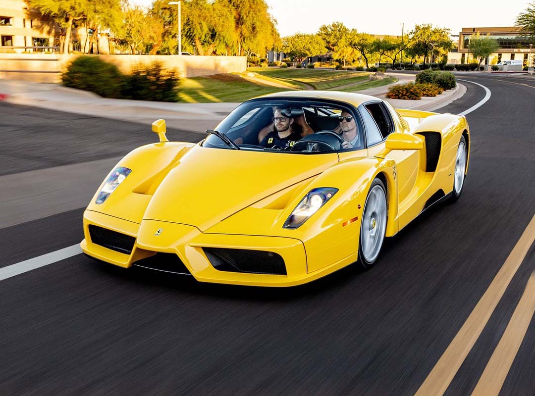 A yellow Ferrari Enzo in striking Giallo Modena, featuring luxurious Cuoio Leather seats, cruises down a multi-lane road lined with trees and buildings on a sunny day with two people inside.