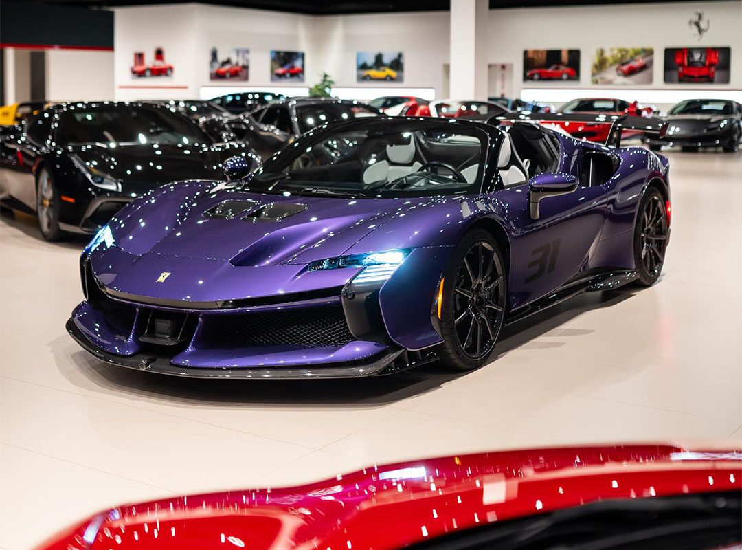 A Violetto Dino Ferrari SF90 Spider is displayed at Ferrari Lake Forest's luxury showroom, surrounded by other high-end vehicles under bright lighting.