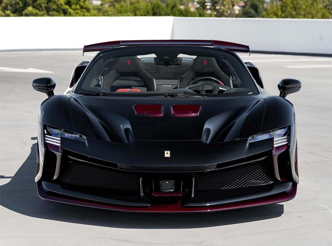 Front view of a black Ferrari Beverly Hills SF90 XX Spider with striking red carbon fiber accents, parked on a rooftop against trees and a white barrier backdrop.