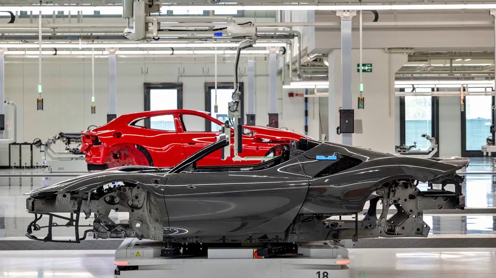 A black EV convertible chassis and a red Ferrari coupe chassis are positioned in a brightly lit automotive factory assembly area.