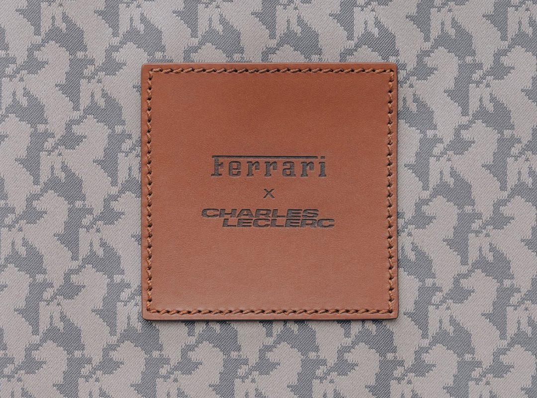 A brown leather patch with "Ferrari x Charles Leclerc" embossed, sewn onto gray fabric with a repeating horse pattern—a touch of luxury perfect for those who follow the latest lifestyle news.